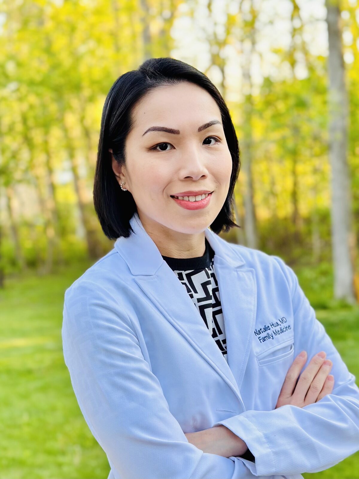 Dr. Natalie Hua smiling outdoors in a white coat embroidered with “Natalie Hua, MD, Family Medicine.”